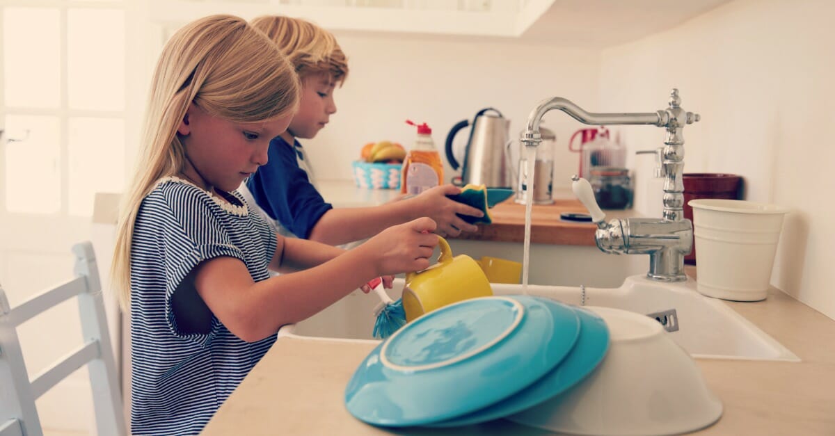 Brother and sister kneeling on chairs to wash up in kitchen